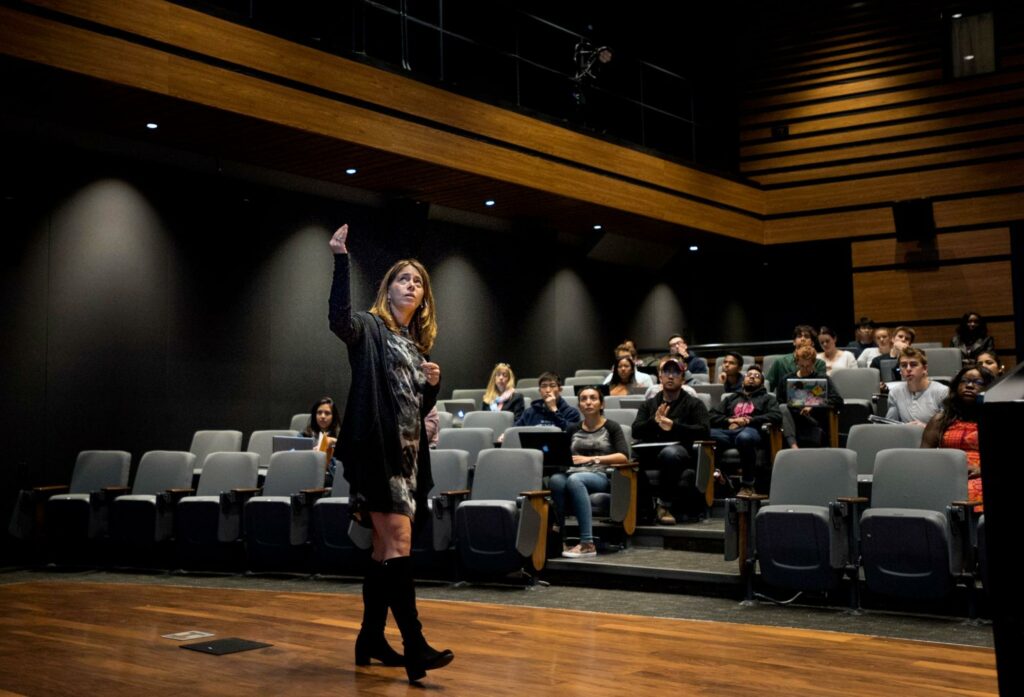 A speaker presents in front of an audience in a lecture hall with seating.