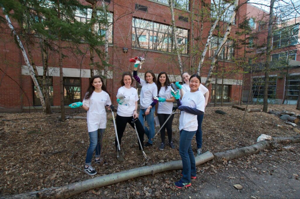 A group of people in white t-shirts are standing outdoors, smiling and holding trash bags and litter pickers while participating in a cleanup activity in a wooded area.